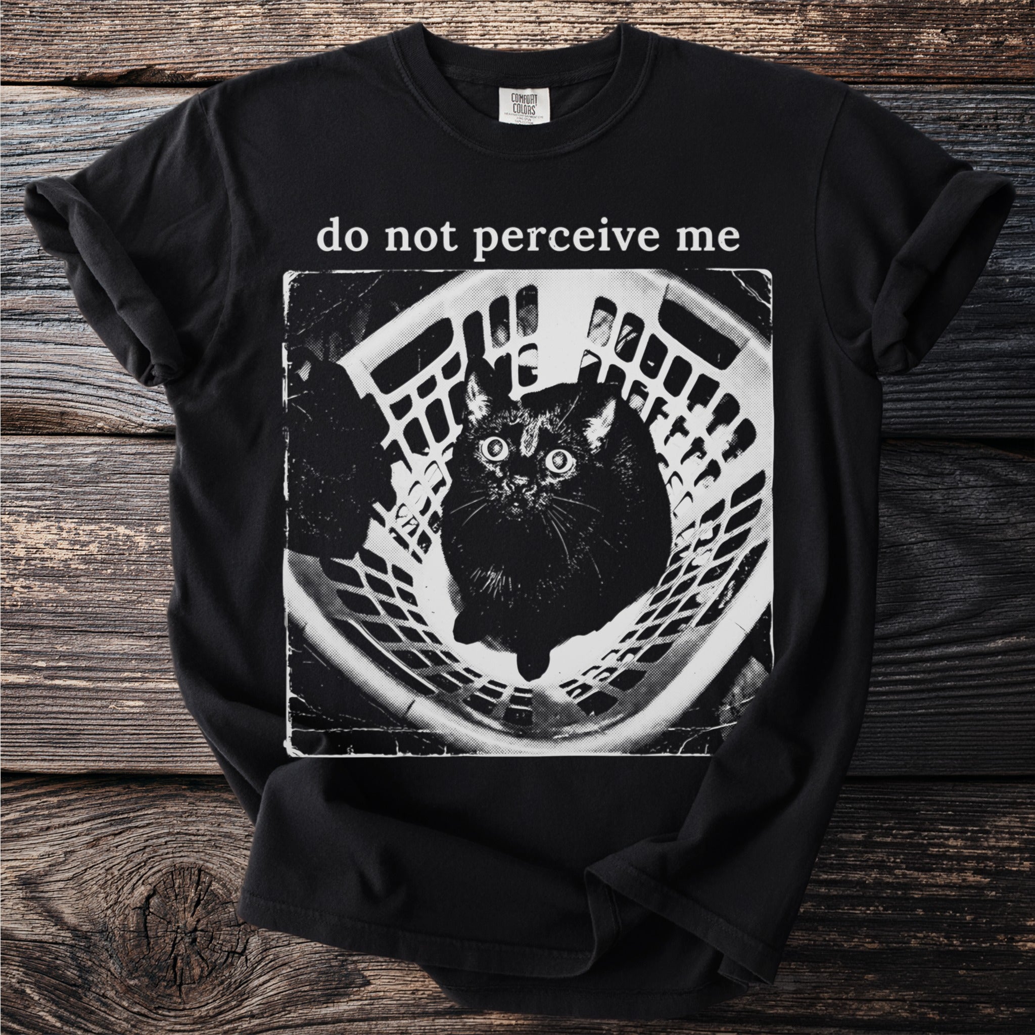 Black t-shirt with "do not perceive me" text and a wide-eyed black cat in a laundry basket.