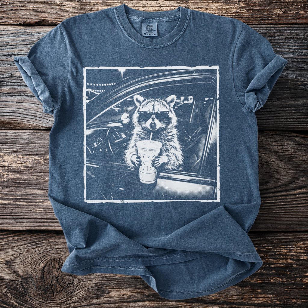 Graphic tee with a raccoon in sunglasses holding a drink inside a car.