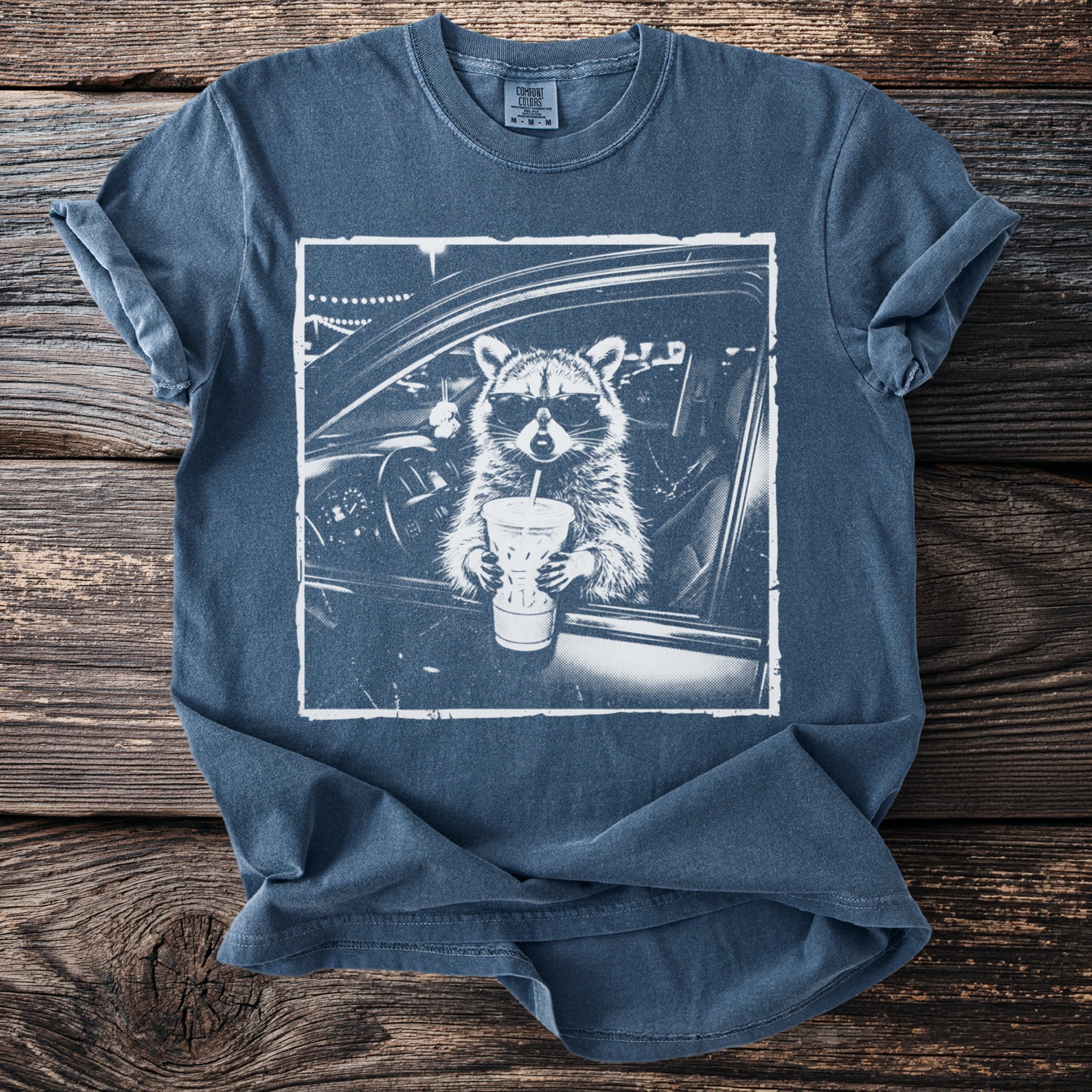 Graphic tee with a raccoon in sunglasses holding a drink inside a car.