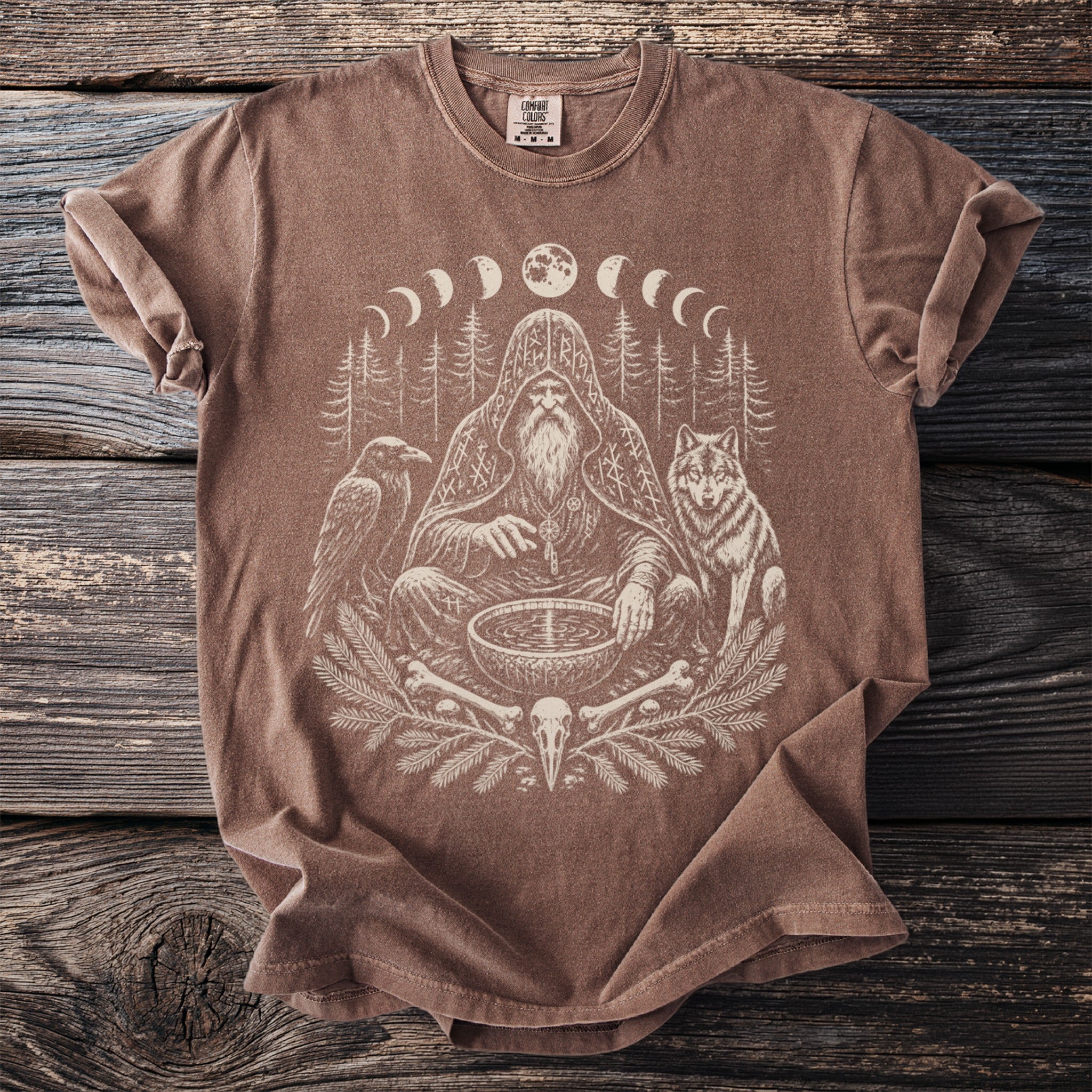 Brown t-shirt with occult graphic of a shaman, raven, wolf, moon phases, and bones.