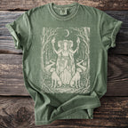 Green t-shirt with a beige illustration of three goddesses, dogs, torches, and a moonlit forest path.