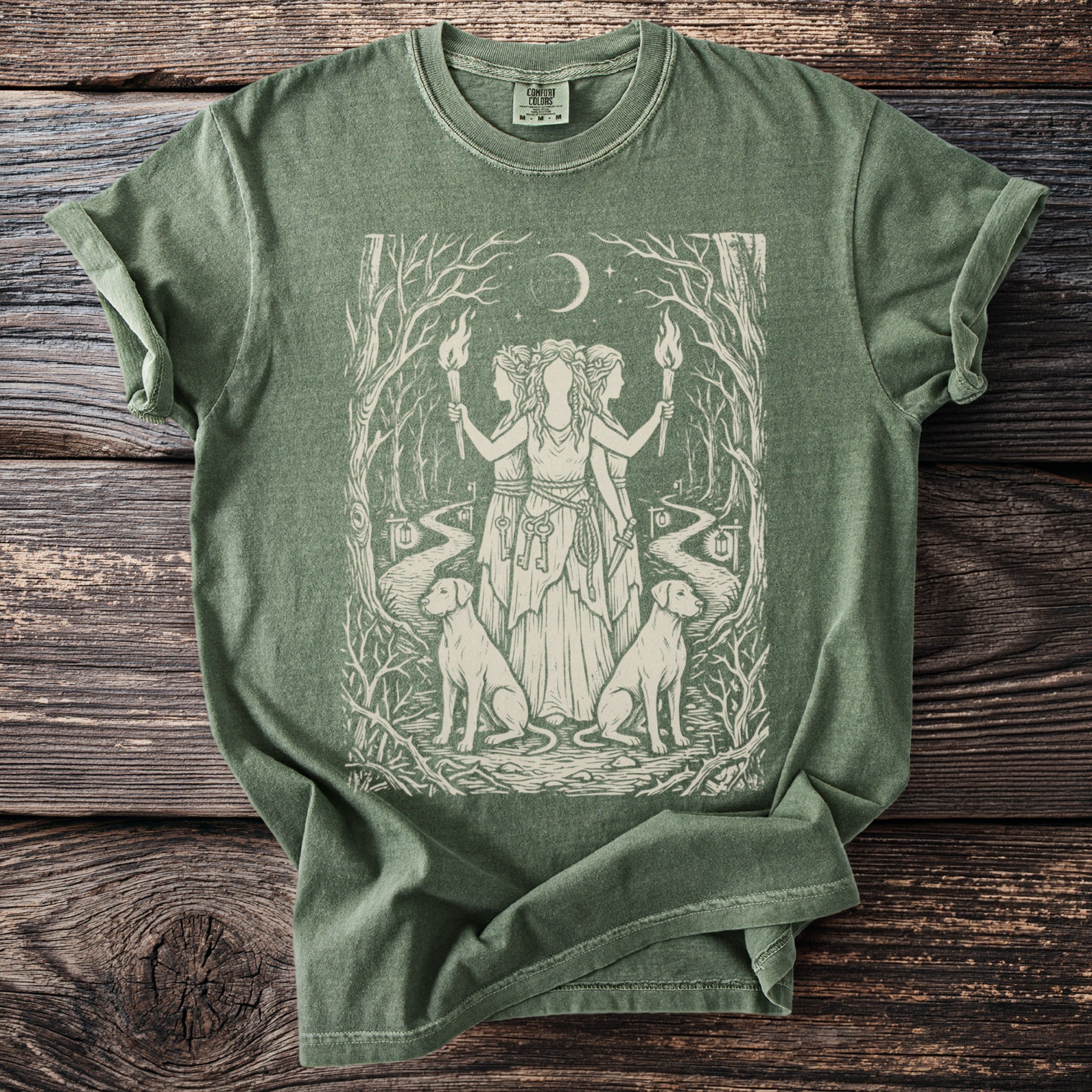 Green t-shirt with a beige illustration of three goddesses, dogs, torches, and a moonlit forest path.