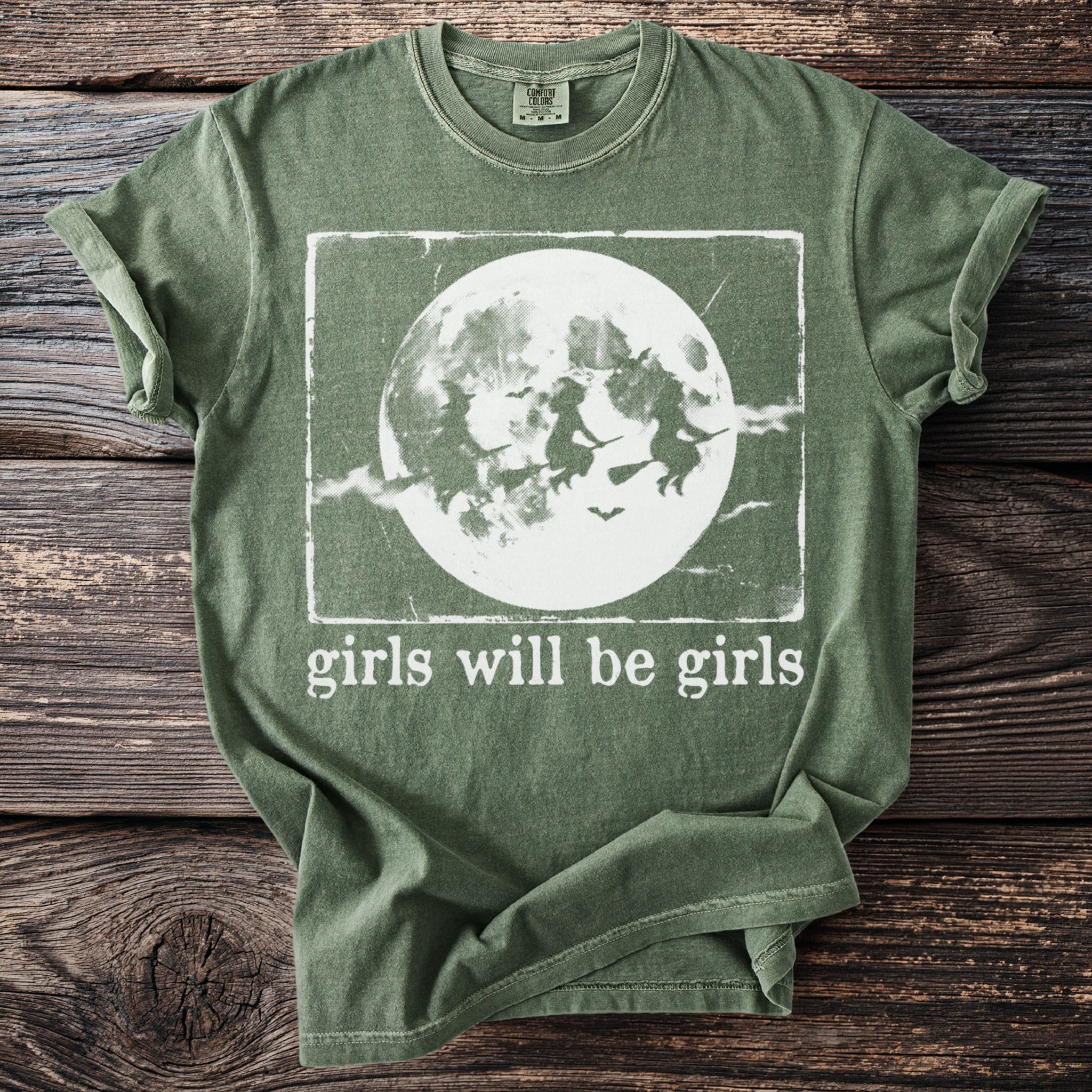 Green t-shirt with witches flying in front of a moon and "girls will be girls" text.