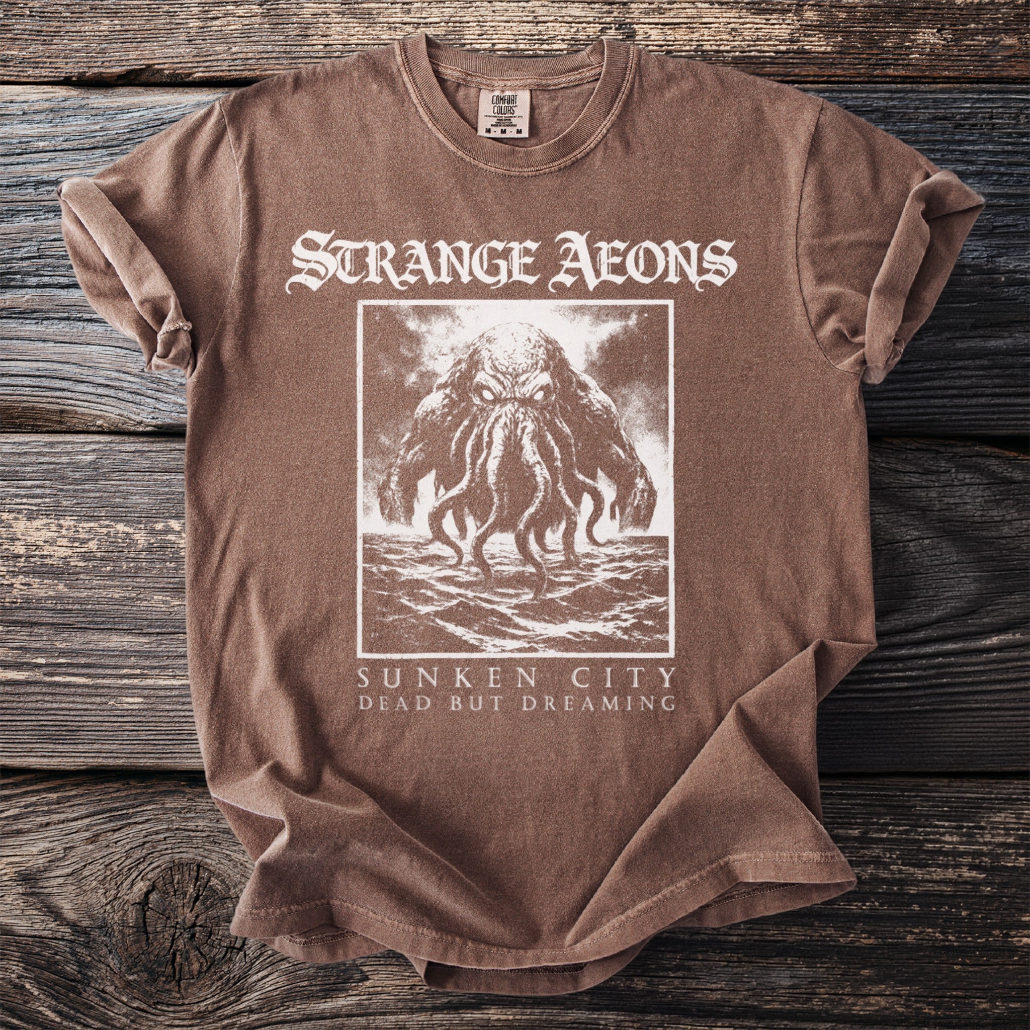 Brown t-shirt with "STRANGE AEONS SUNKEN CITY DEAD BUT DREAMING" and tentacled creature graphic.