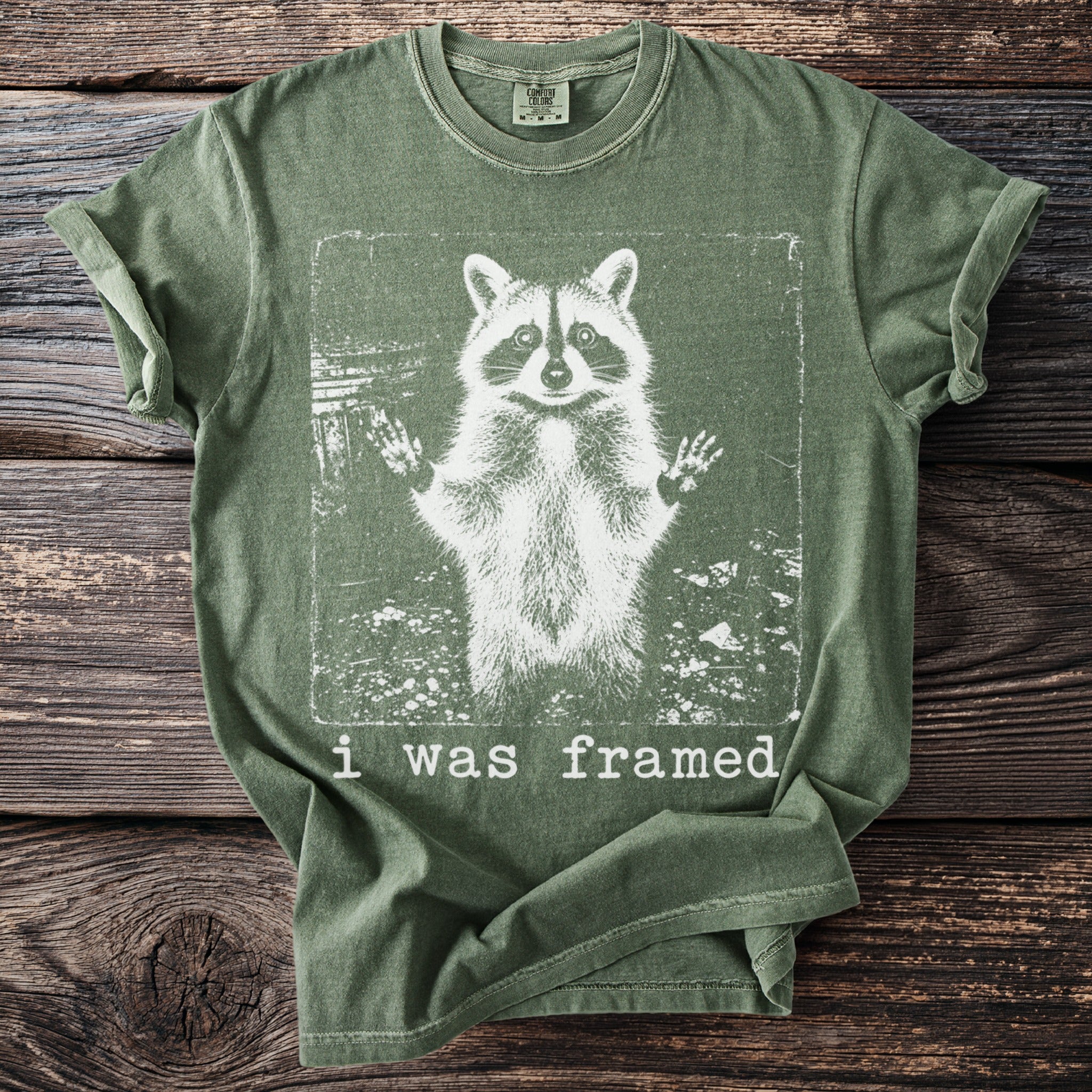 Green t-shirt with a distressed print of a raccoon with raised paws and the text "i was framed."