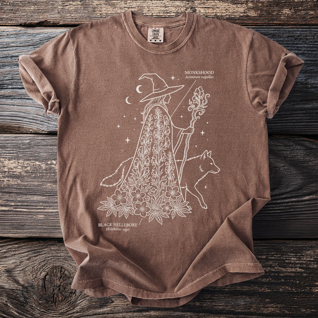 Brown t-shirt with line art of a witch, fox, monkshood, and black hellebore.