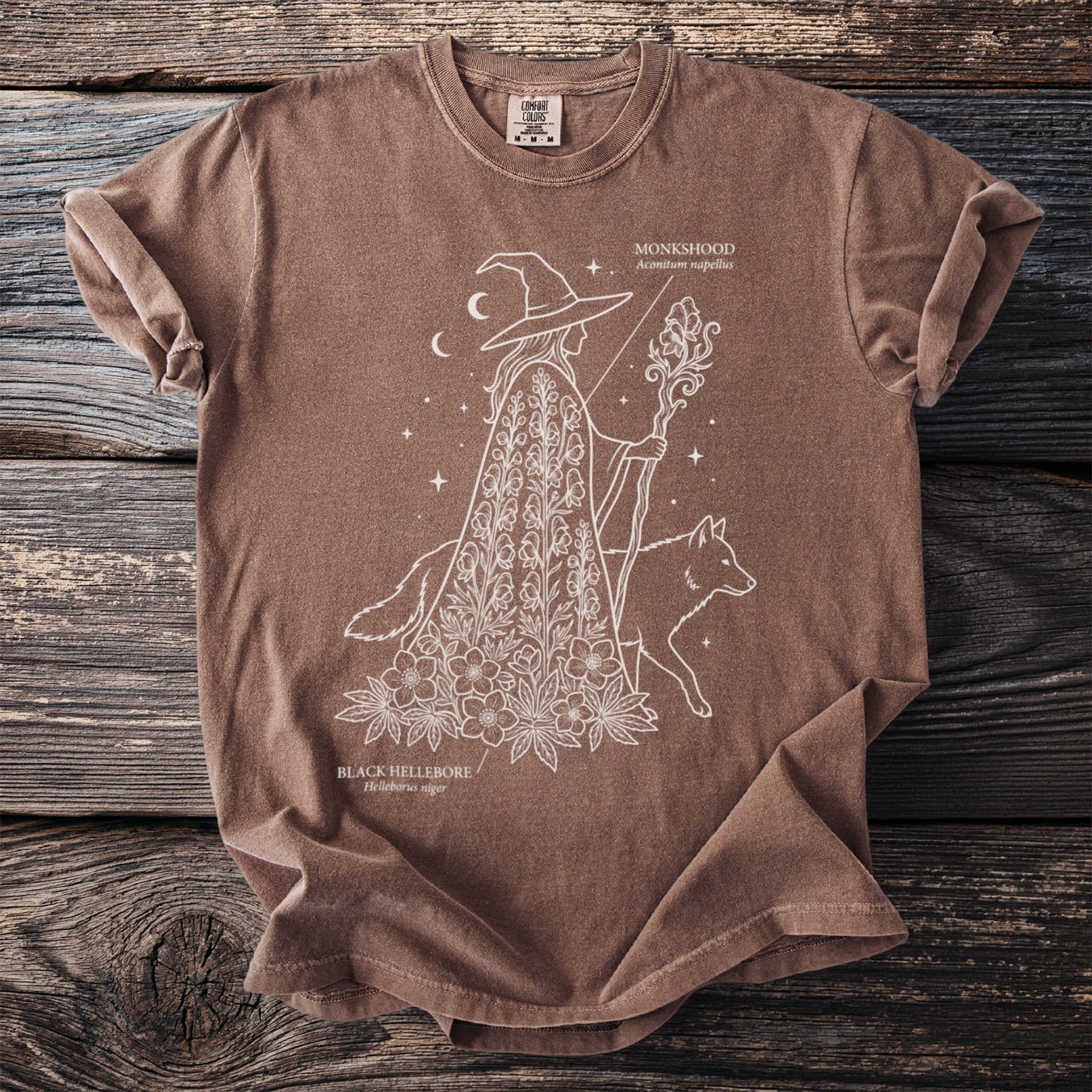 Brown t-shirt with line art of a witch, fox, monkshood, and black hellebore.