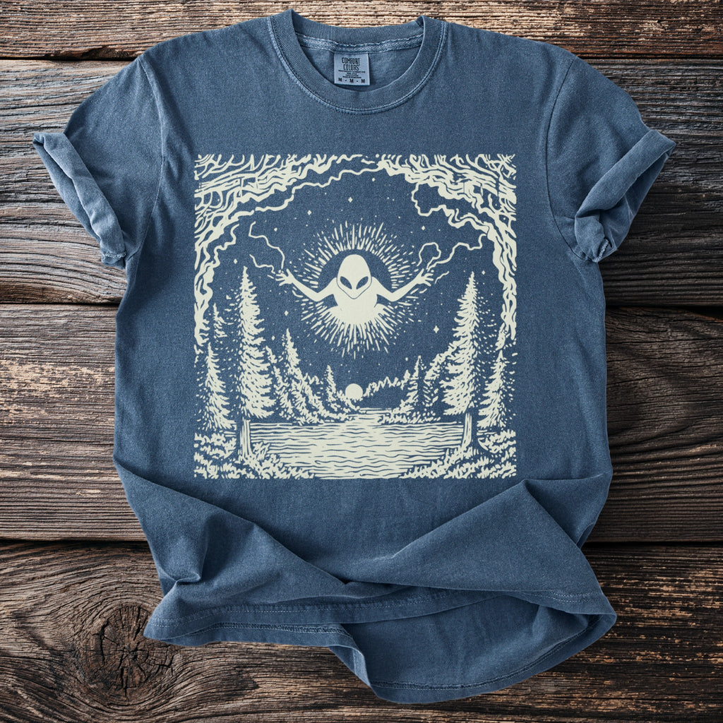 T-shirt with a cream-colored graphic of an alien in a forest scene with a lake.