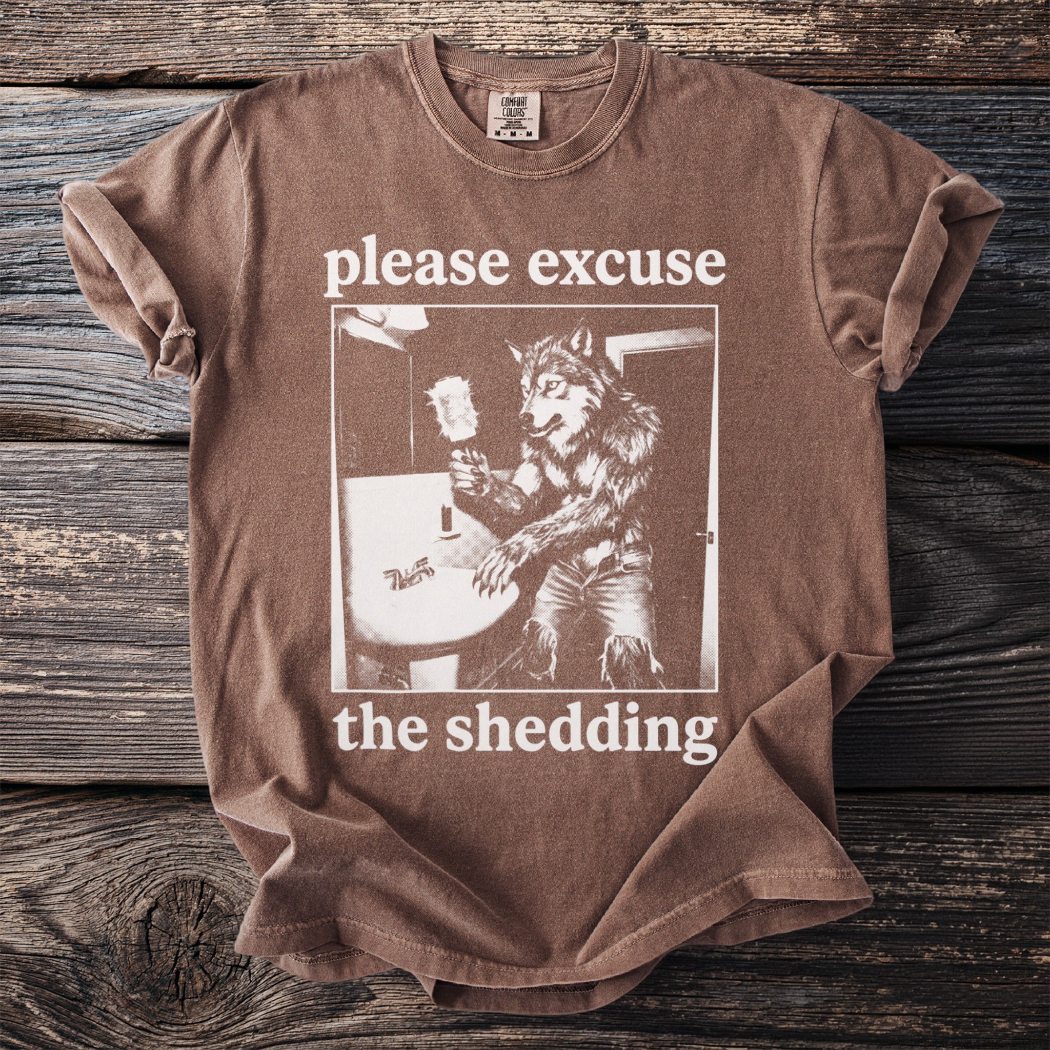 Brown t-shirt with graphic of a werewolf by a sink, text says "please excuse the shedding".