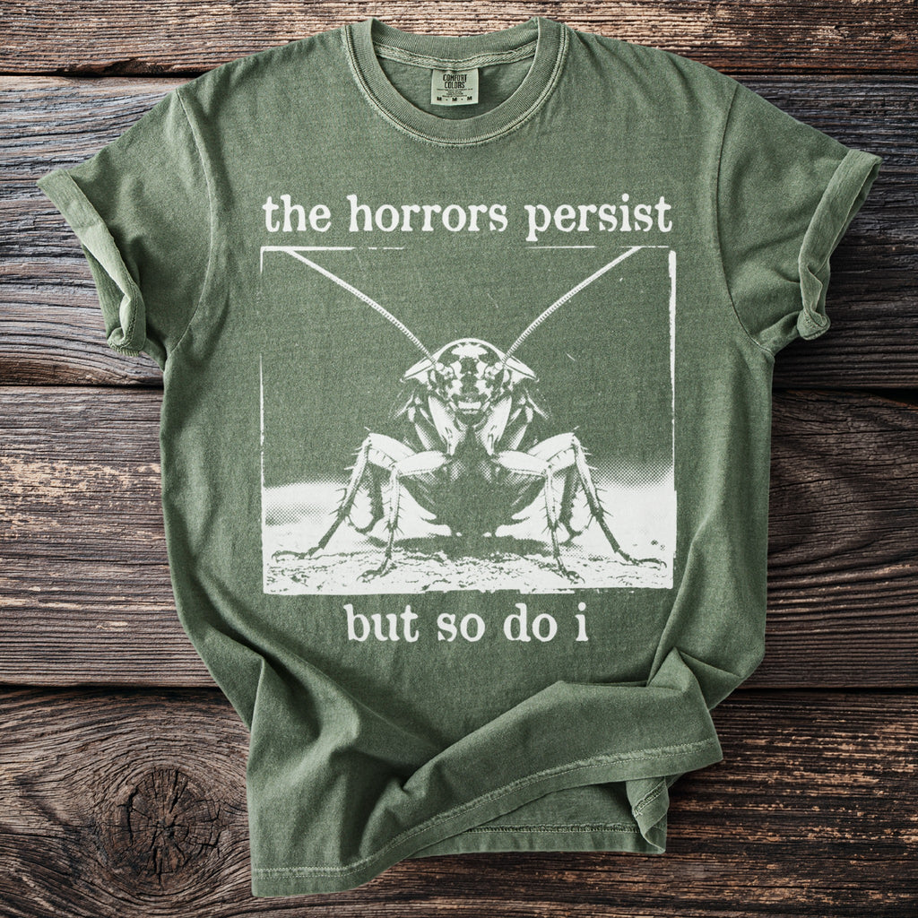 Green t-shirt with cockratch graphic and text "the horrors persist but so do i".