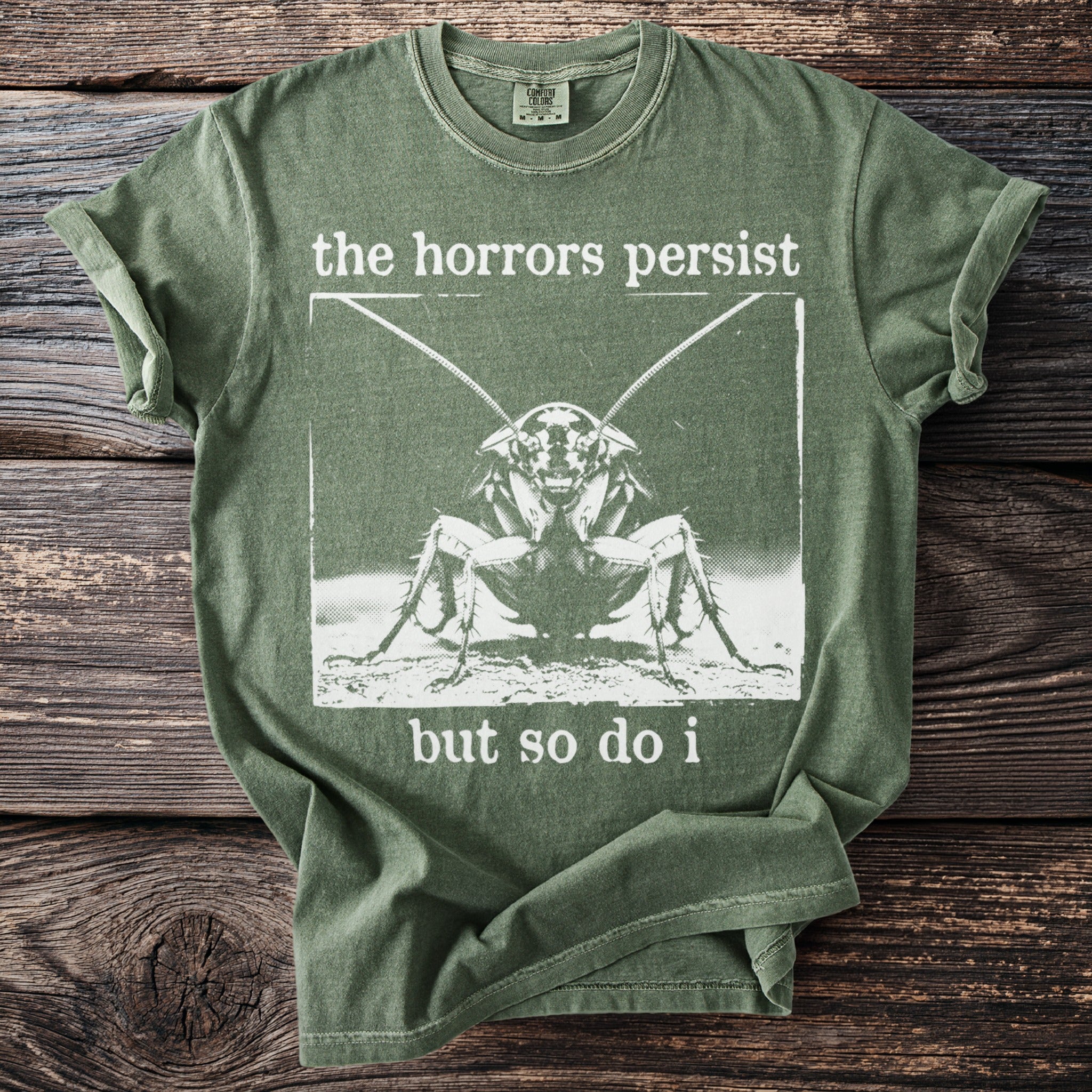 Green t-shirt with cockratch graphic and text "the horrors persist but so do i".