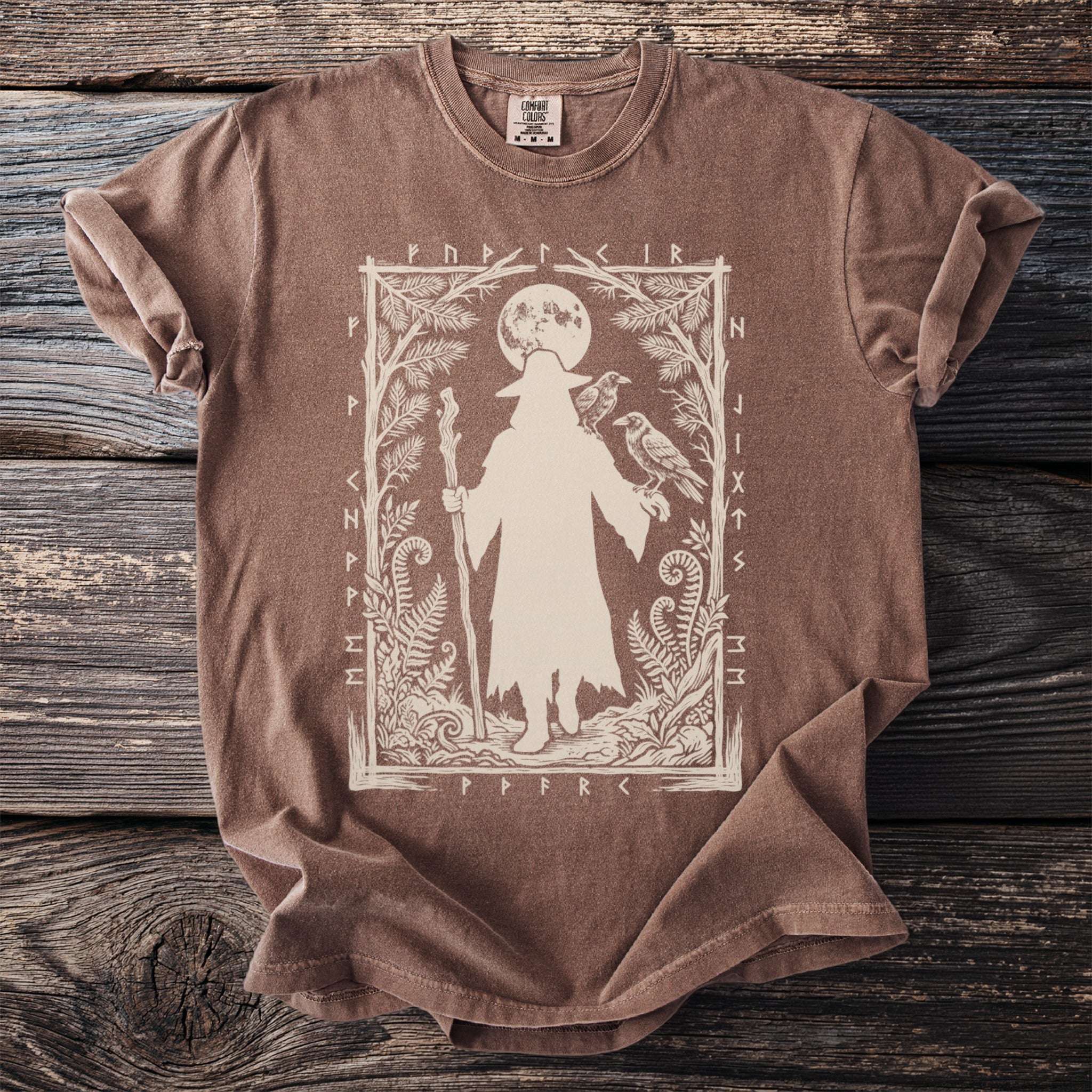 Brown t-shirt with a silhouette of a figure holding a staff and two ravens, surrounded by foliage and runes.