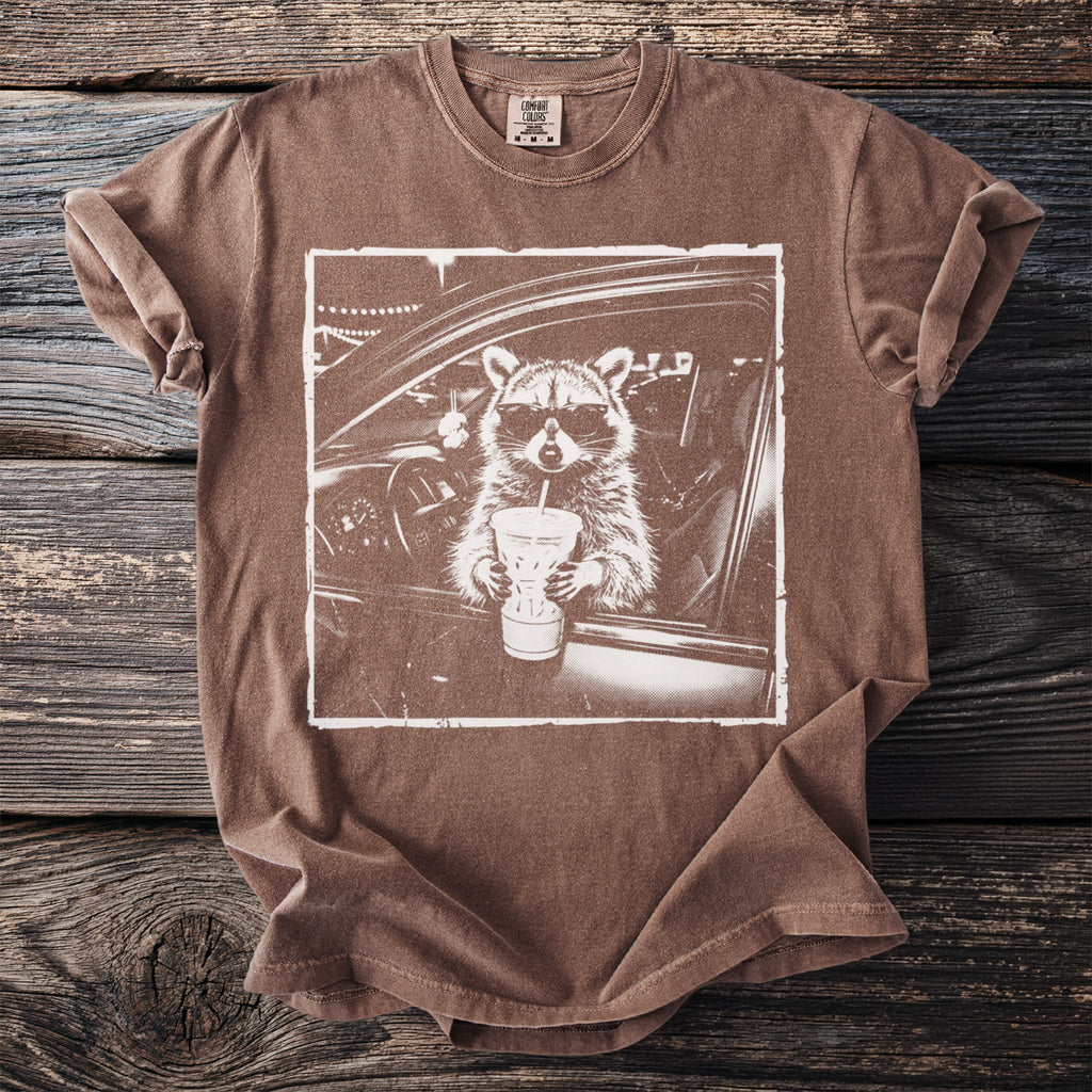 Brown t-shirt with a graphic of a raccoon wearing sunglasses drinking from a cup.