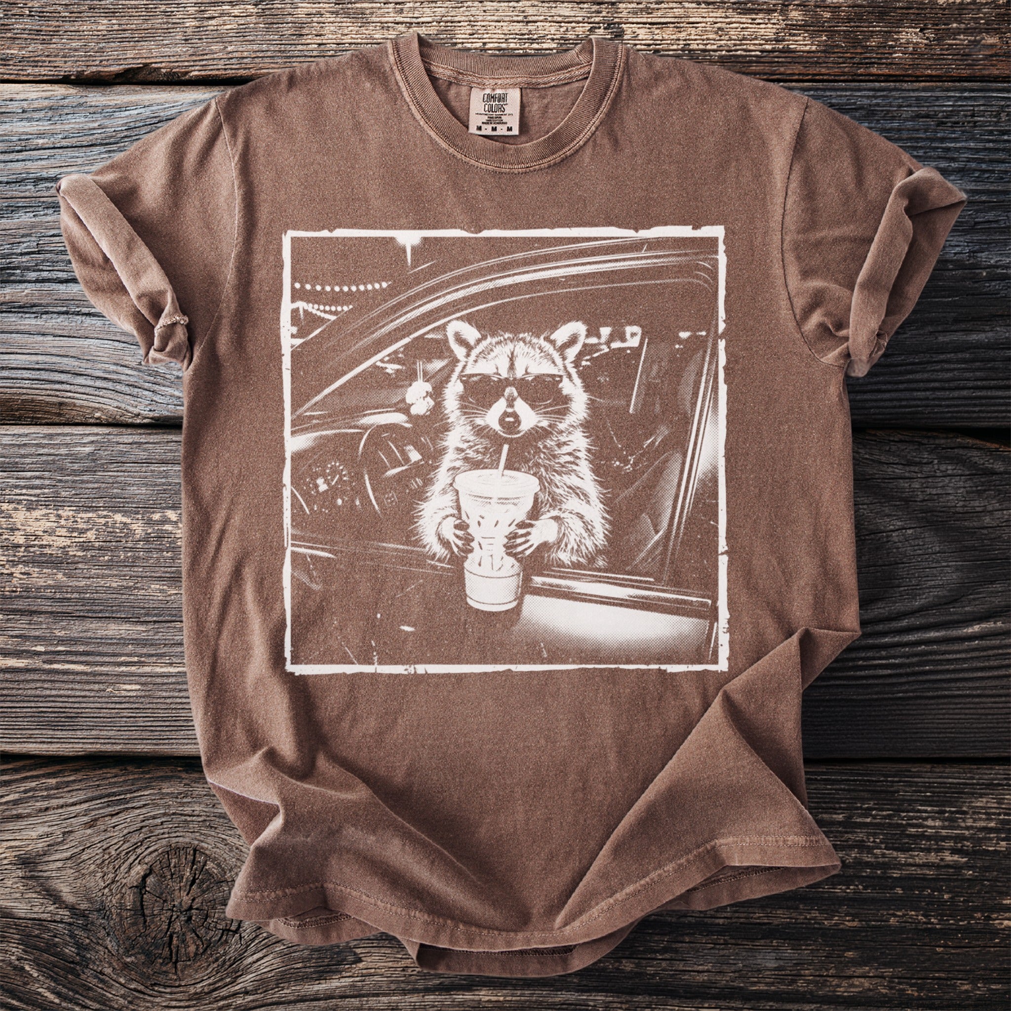 Brown t-shirt with a graphic of a raccoon wearing sunglasses drinking from a cup.