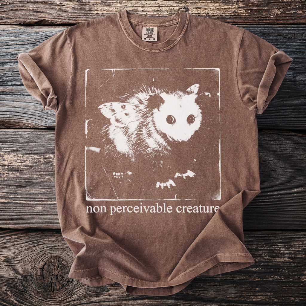 Brown t-shirt with white graphic of an opossum and moth and text "non perceivable creature".