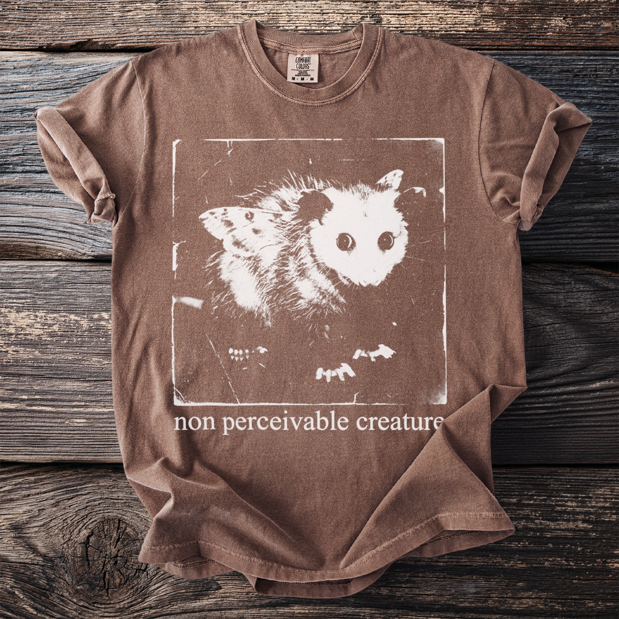 Brown t-shirt with white graphic of an opossum and moth and text "non perceivable creature".