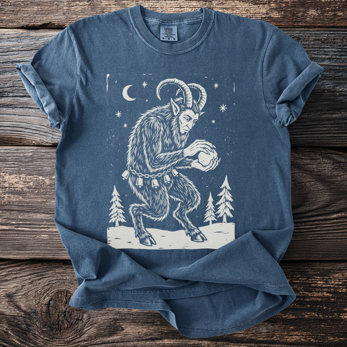 Denim blue t-shirt with a cream folk-cut print: Krampus crouches among snowy pines, packing a snowball between his clawed hands.