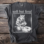 Dark grey t-shirt with "soft but feral" and a werewolf looking into a box with a kitten.