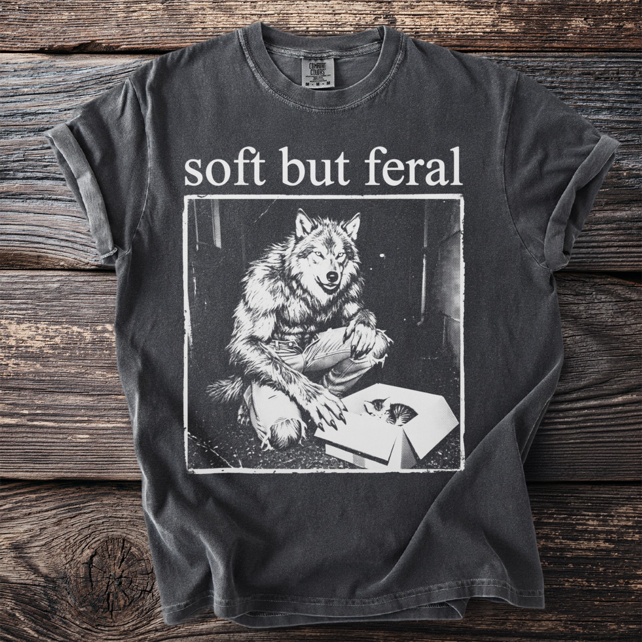Dark grey t-shirt with "soft but feral" and a werewolf looking into a box with a kitten.