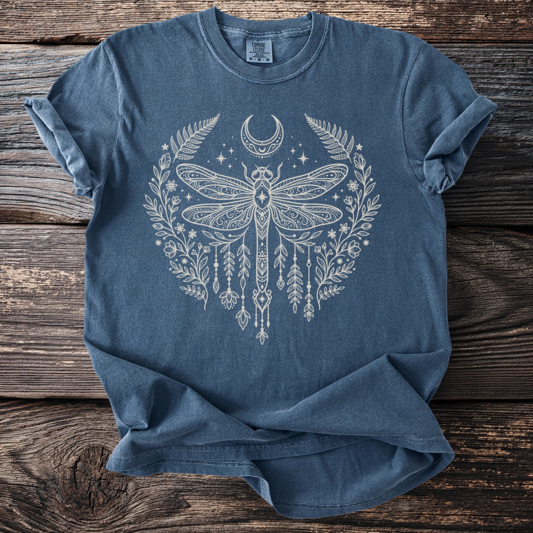 Blue t-shirt with white celestial dragonfly and floral design on a wooden background.