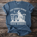 T-shirt with "God Forbid Women Have Hobbies" text featuring witch watering plants at night.