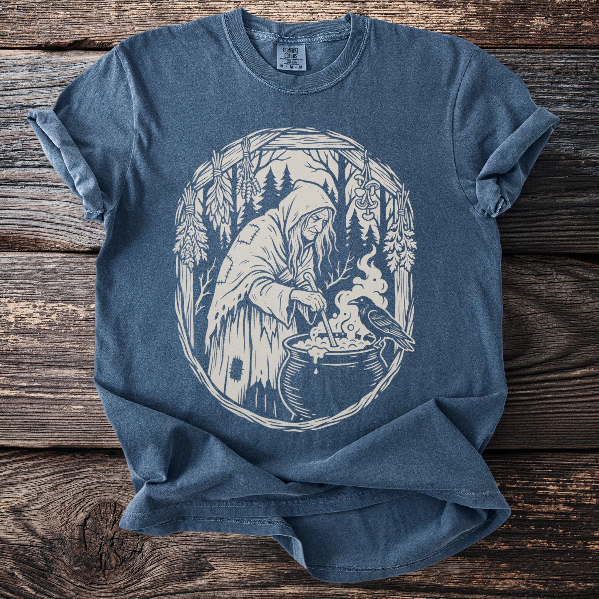 T-shirt with cream design of a witch stirring cauldron with a crow beside it.