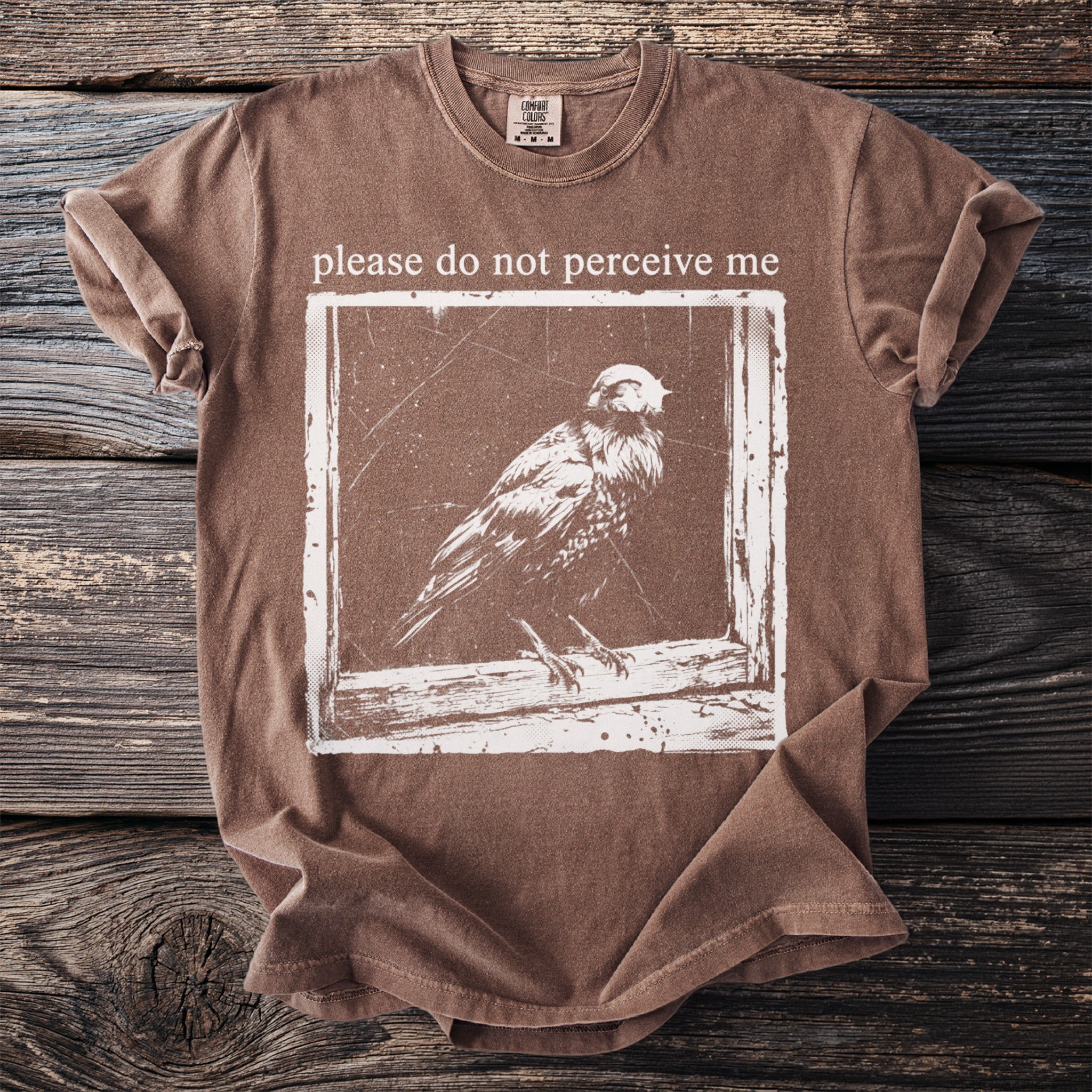 Brown t-shirt with white graphic of a bird and text "please do not perceive me."