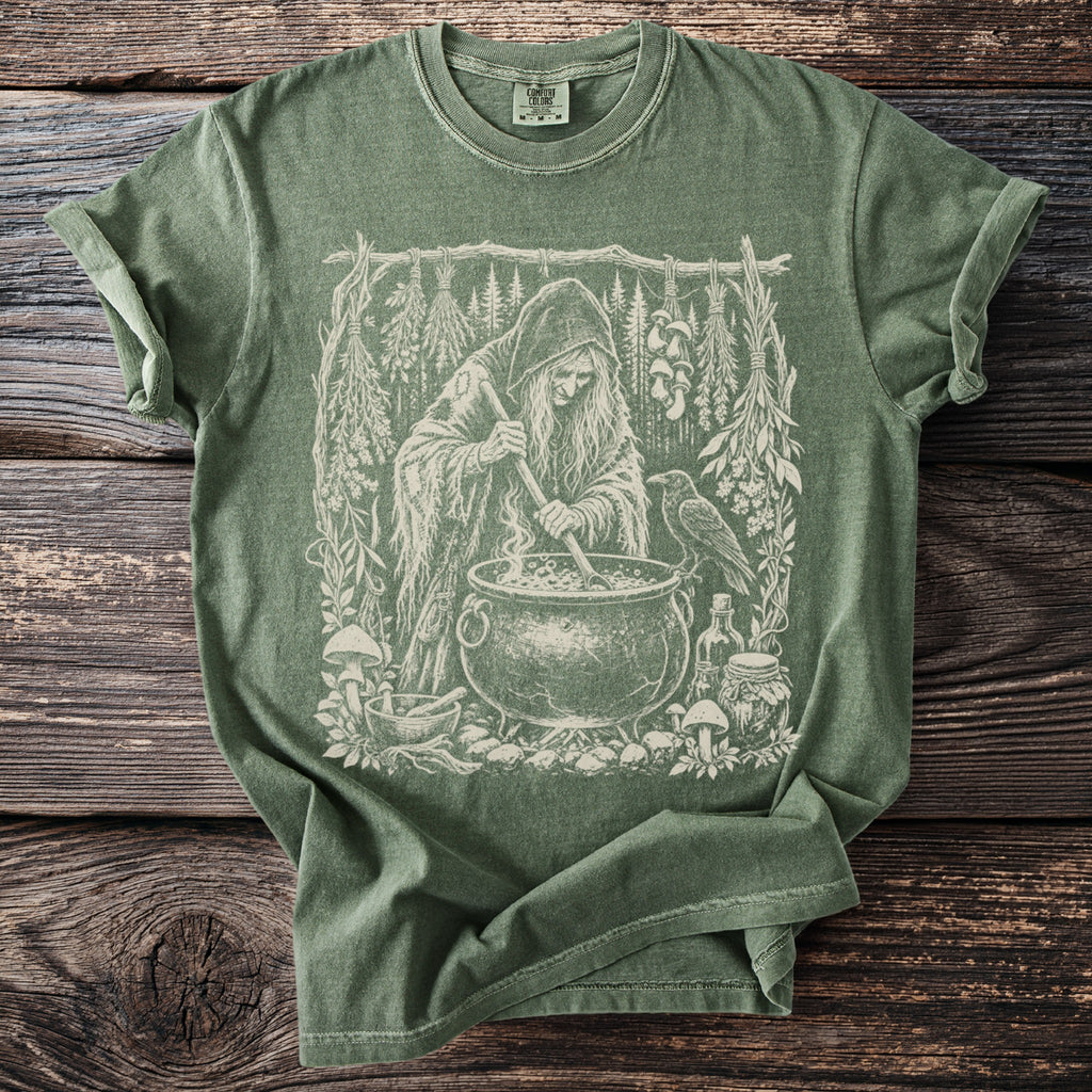 Green t-shirt with a white ink drawing of a witch tending a cauldron with a raven.