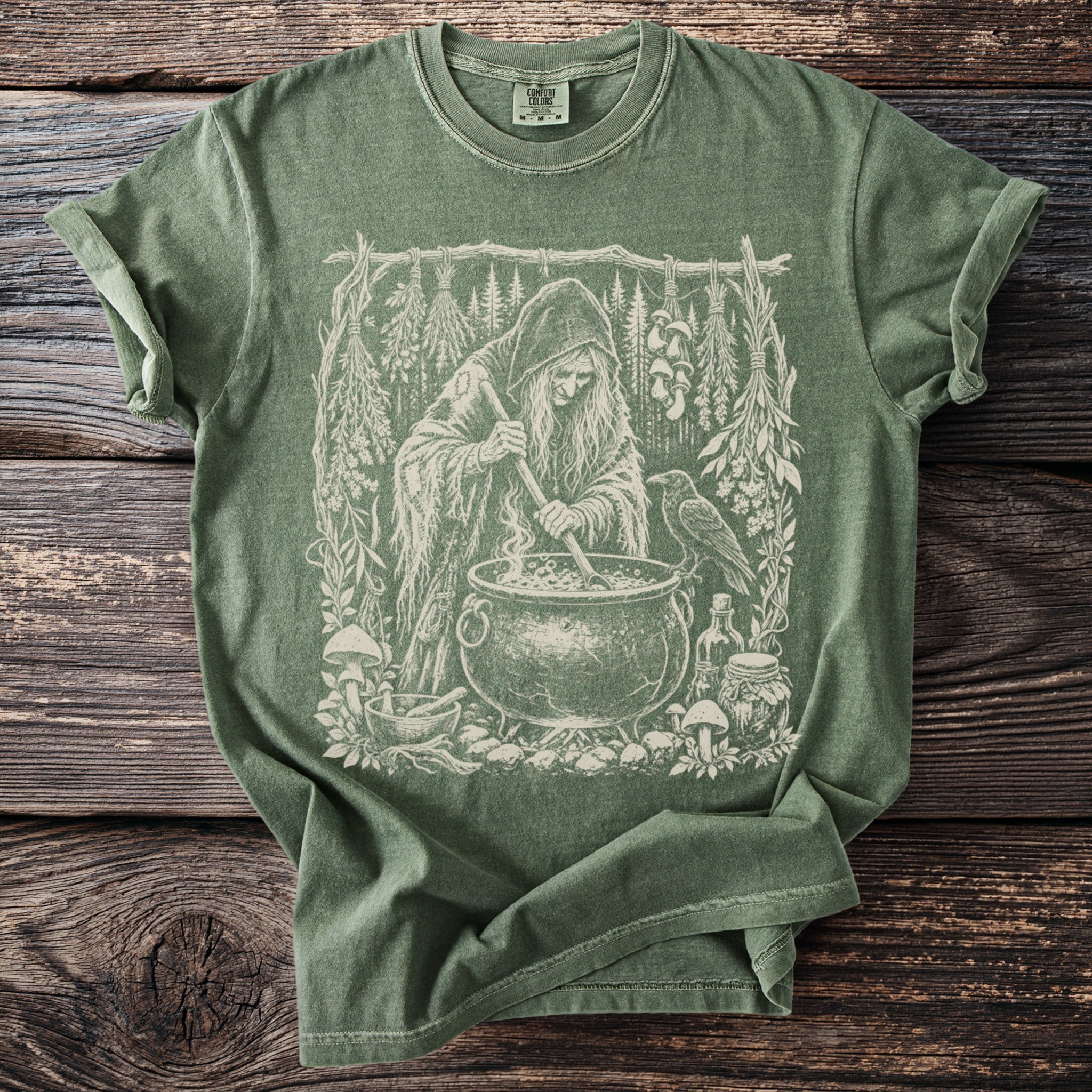Green t-shirt with a white ink drawing of a witch tending a cauldron with a raven.