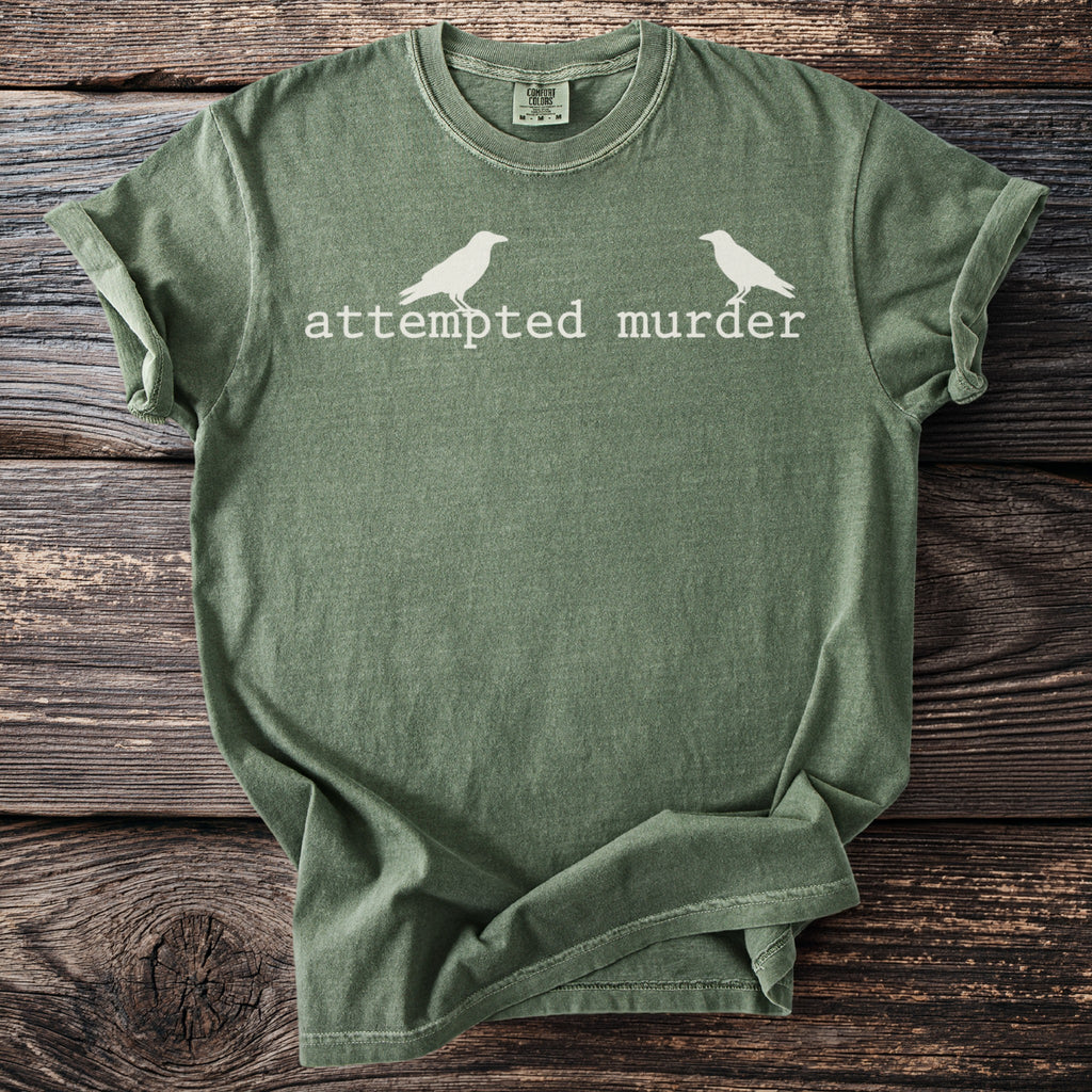 Green t-shirt with white text "attempted murder" and two bird silhouettes.