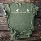 Green t-shirt with white text "attempted murder" and two bird silhouettes.