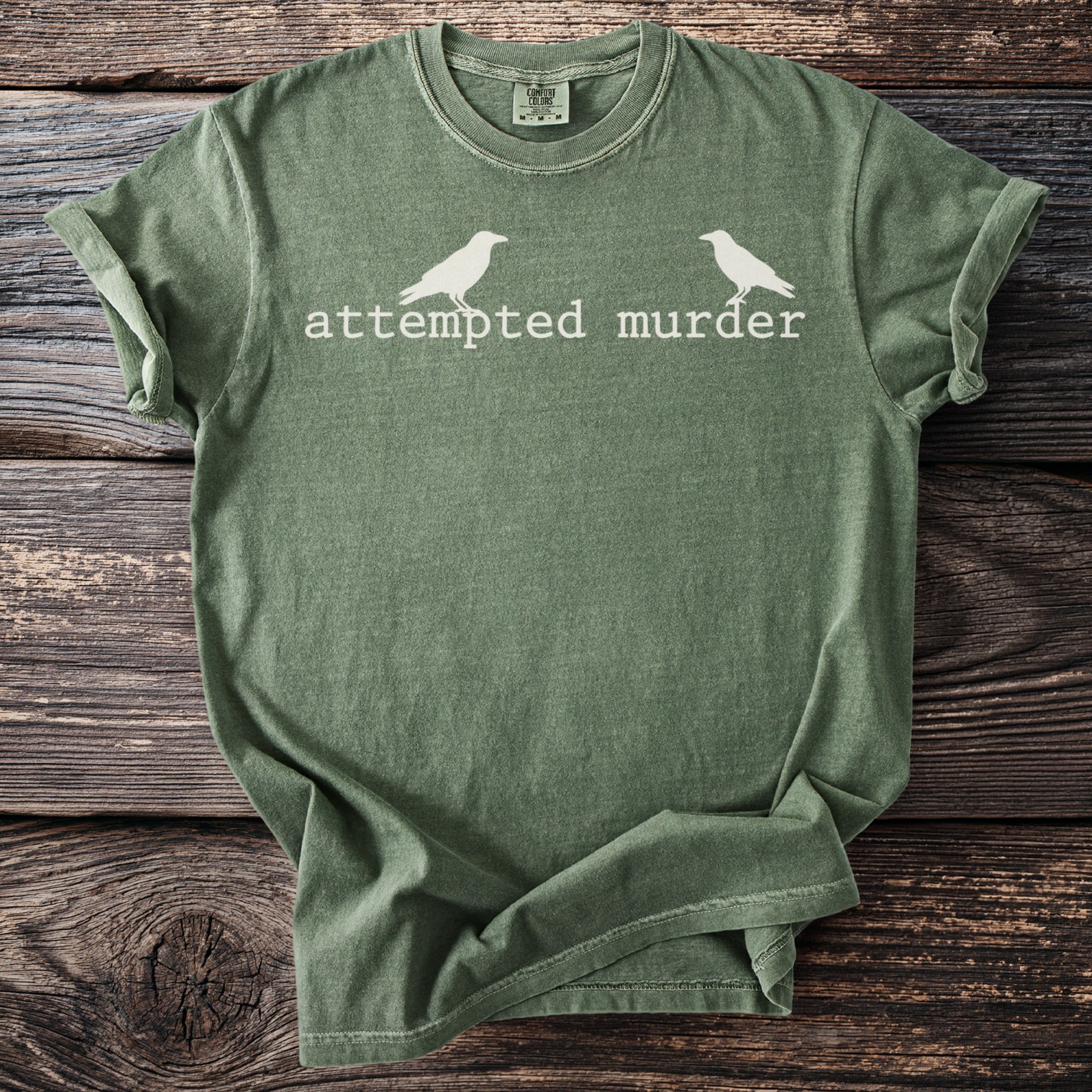 Green t-shirt with white text "attempted murder" and two bird silhouettes.