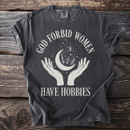 Dark t-shirt with white text and graphics: "GOD FORBID WOMEN HAVE HOBBIES"
