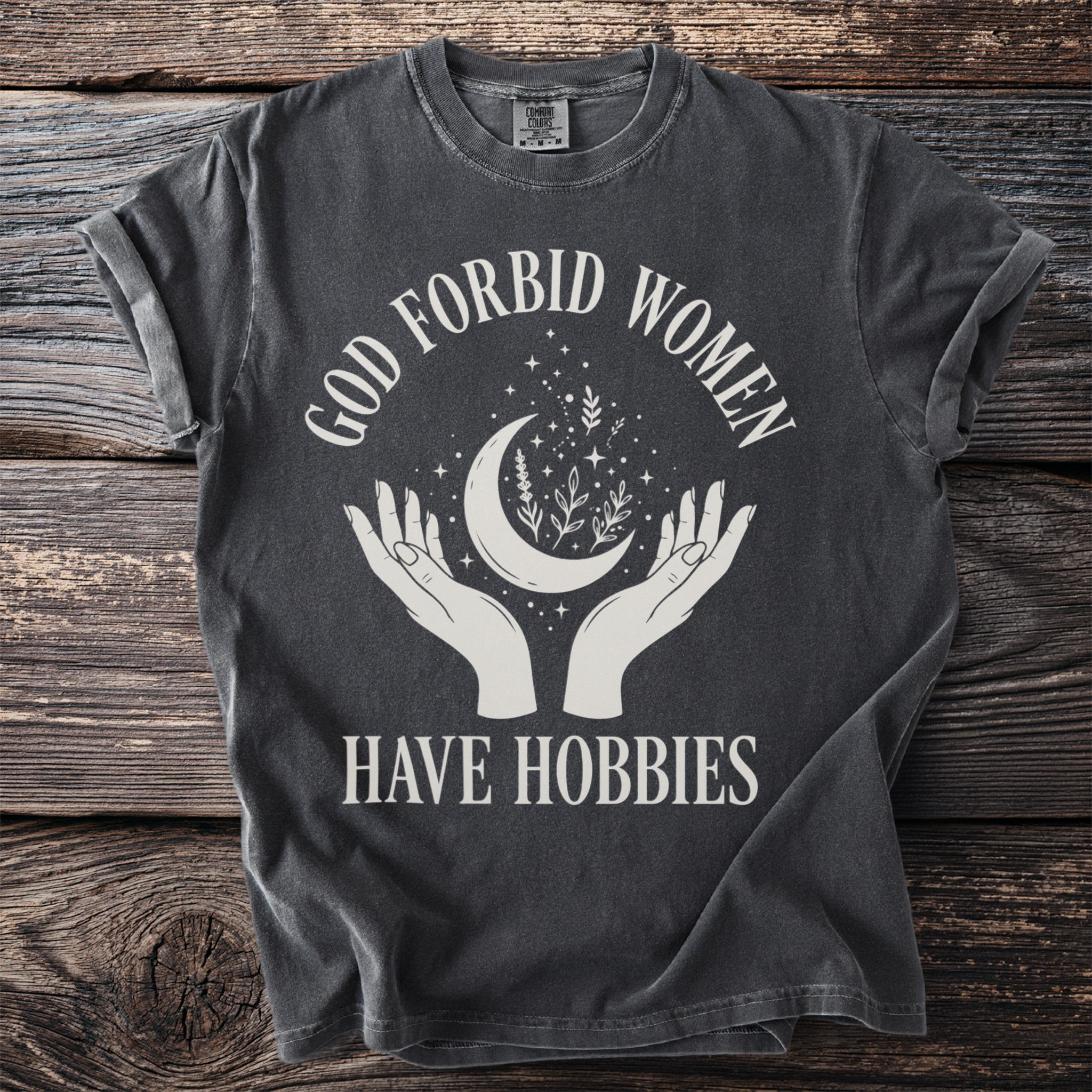 Dark t-shirt with white text and graphics: "GOD FORBID WOMEN HAVE HOBBIES"