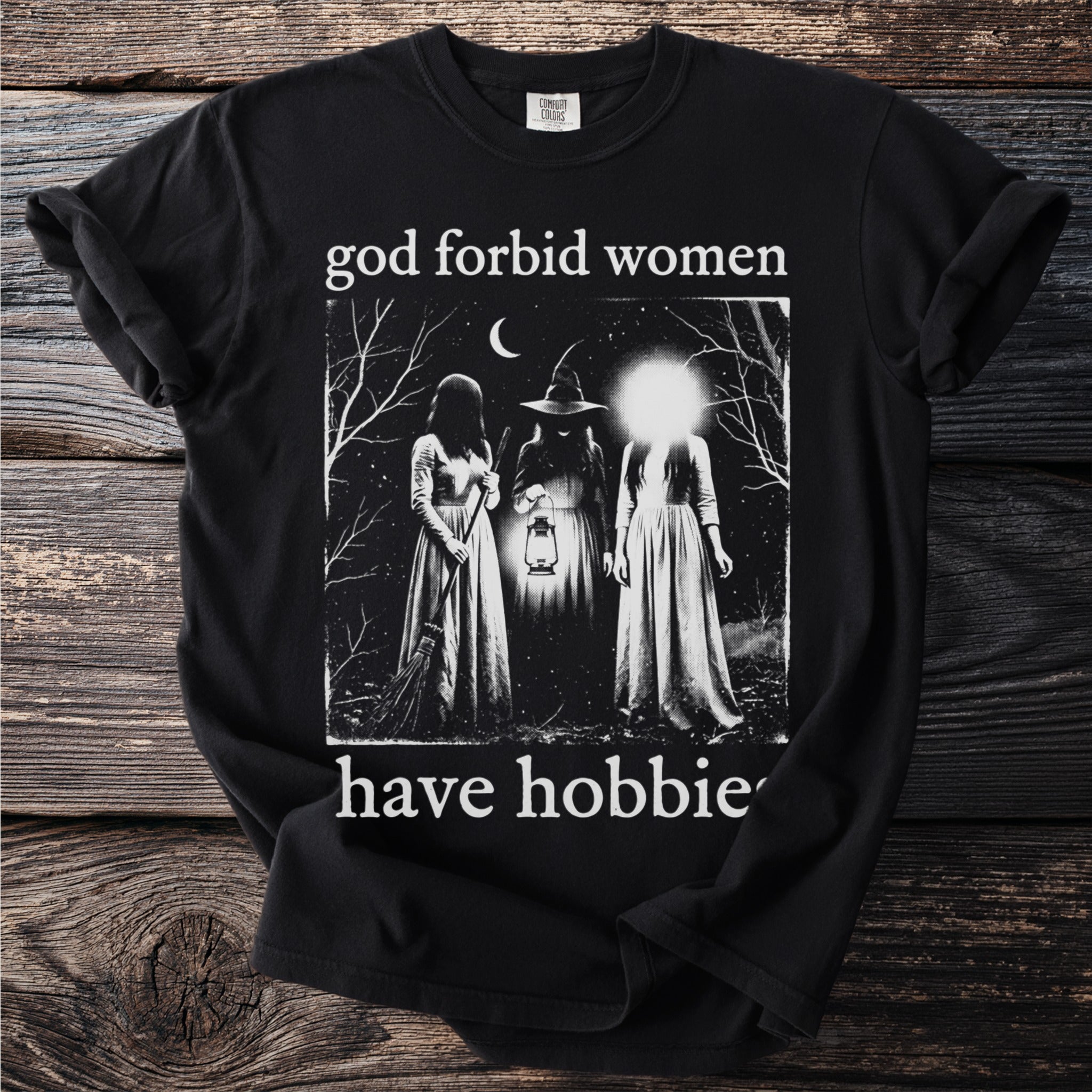 Black t-shirt with "god forbid women have hobbies" and three figures in a forest.