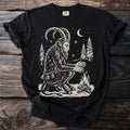 Black t-shirt with a cream folk-cut print: Krampus sits in the snow among tall pines, warming his clawed hands over a small fire.