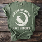 Green t-shirt with white text "GOD FORBID WOMEN HAVE HOBBIES" and a raven on a moon.