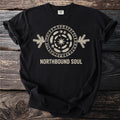 Black t-shirt with a cream circular design of runes, feathers, paw prints, and a star. Text reads "NORTHBOUND SOUL."