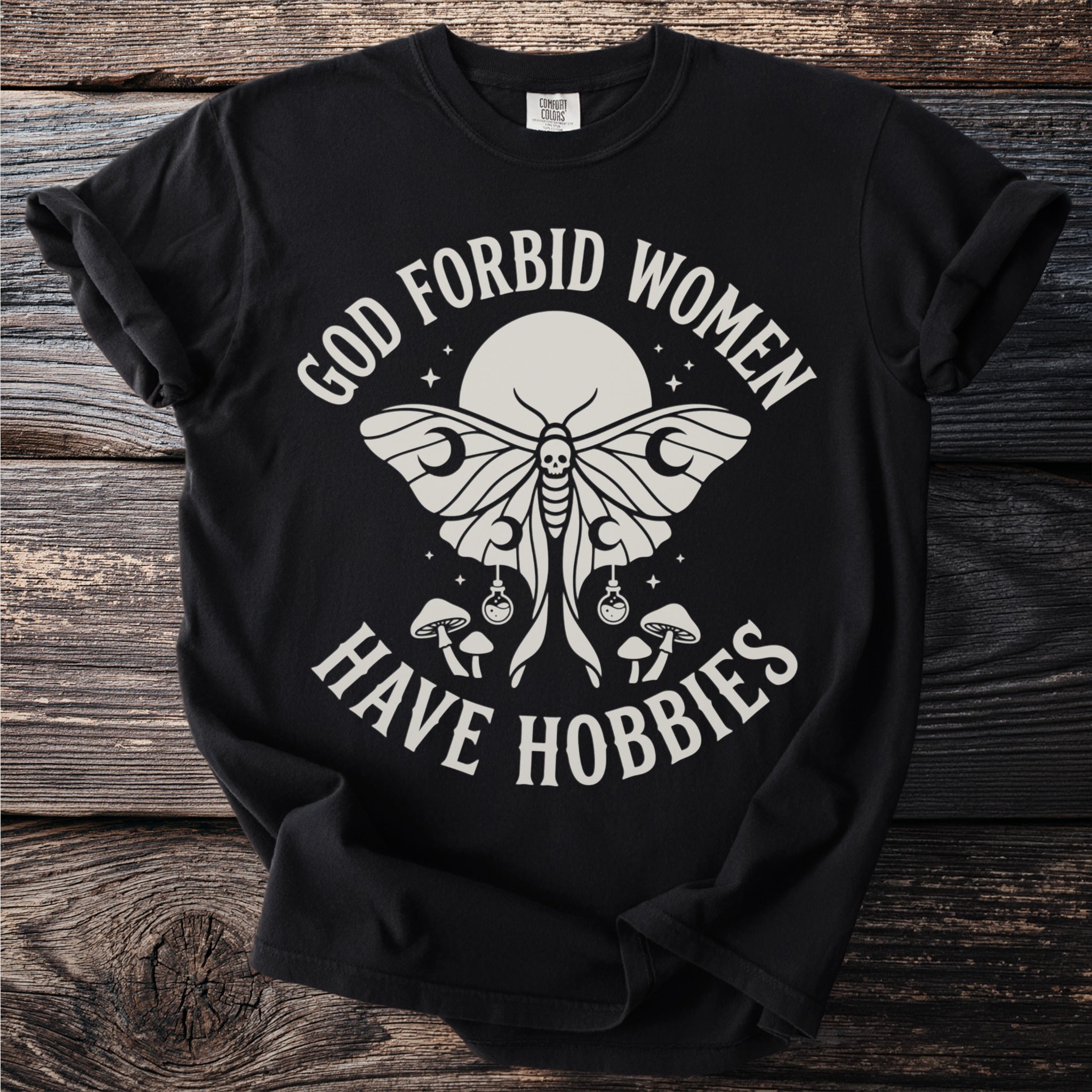 Black Comfort Colors t-shirt with "God Forbid Women Have Hobbies" text and moth graphic.