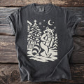 Pepper gray t-shirt with a cream folk-cut print: a raccoon walks away from a decorated tree in the snow, a wrapped present under one arm.