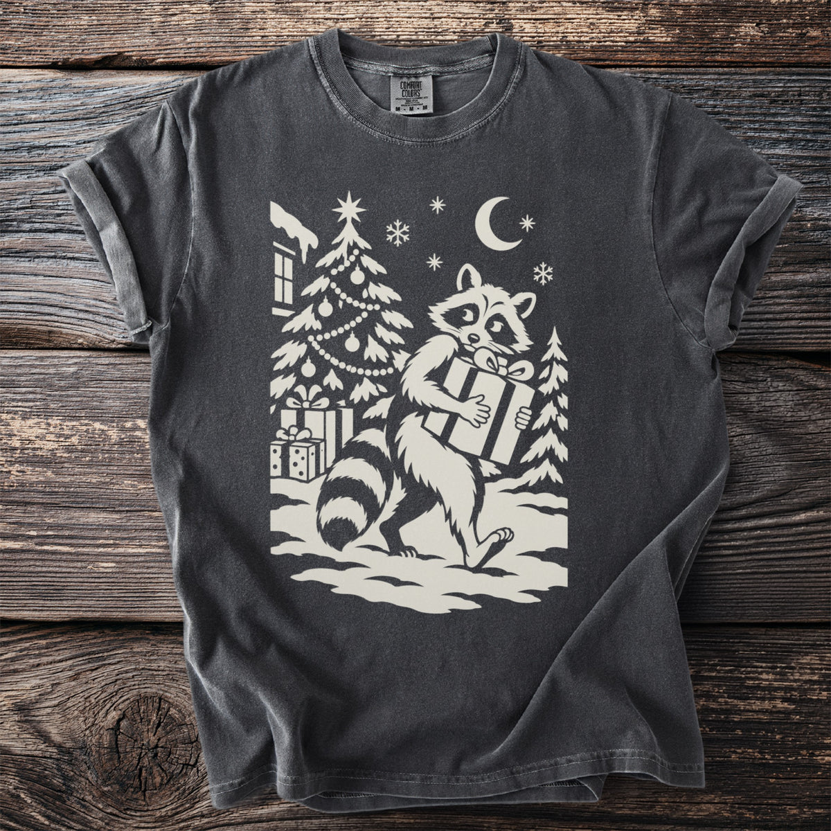 Pepper gray t-shirt with a cream folk-cut print: a raccoon walks away from a decorated tree in the snow, a wrapped present under one arm.