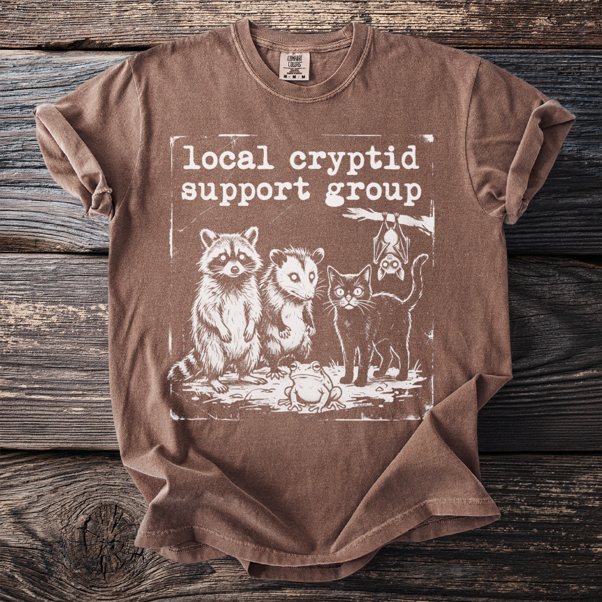 Brown t-shirt with "local cryptid support group" text and illustrations of a raccoon, opossum, cat, frog, and bat.