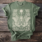 Green t-shirt with a cream graphic of a moth-woman, celestial elements, roses, and mushrooms.