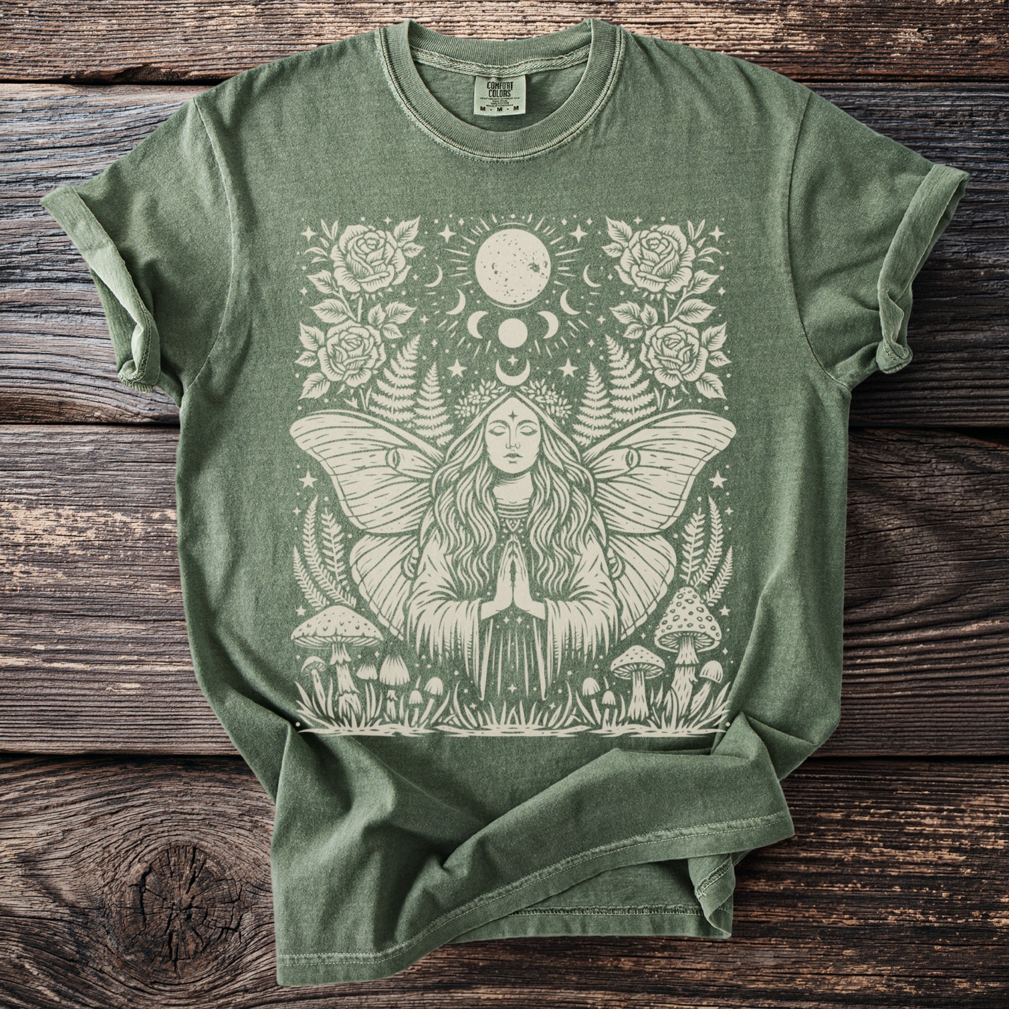 Green t-shirt with a cream graphic of a moth-woman, celestial elements, roses, and mushrooms.