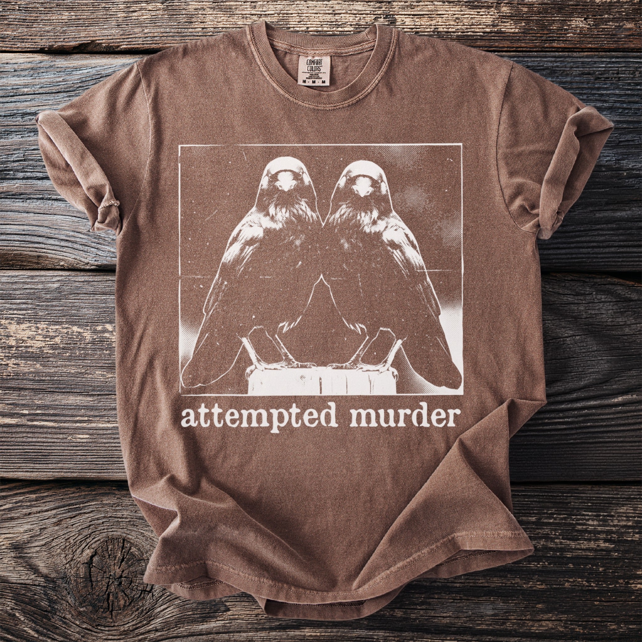 Brown t-shirt with white graphic of two birds and the text "attempted murder".