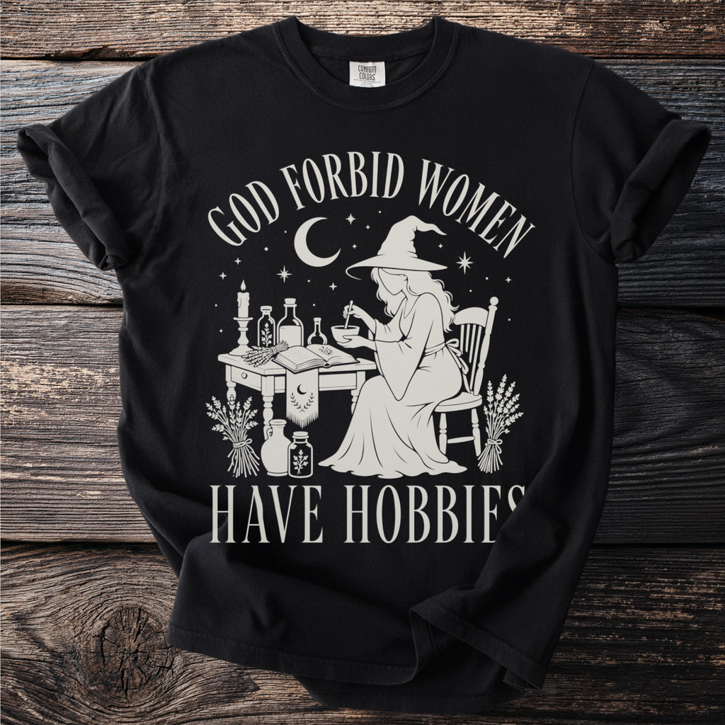 Black t-shirt with cream graphic: "God Forbid Women Have Hobbies"; witch making potions.