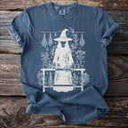 T-shirt with white graphic of a witch, potion bottles, herbs, and celestial elements.