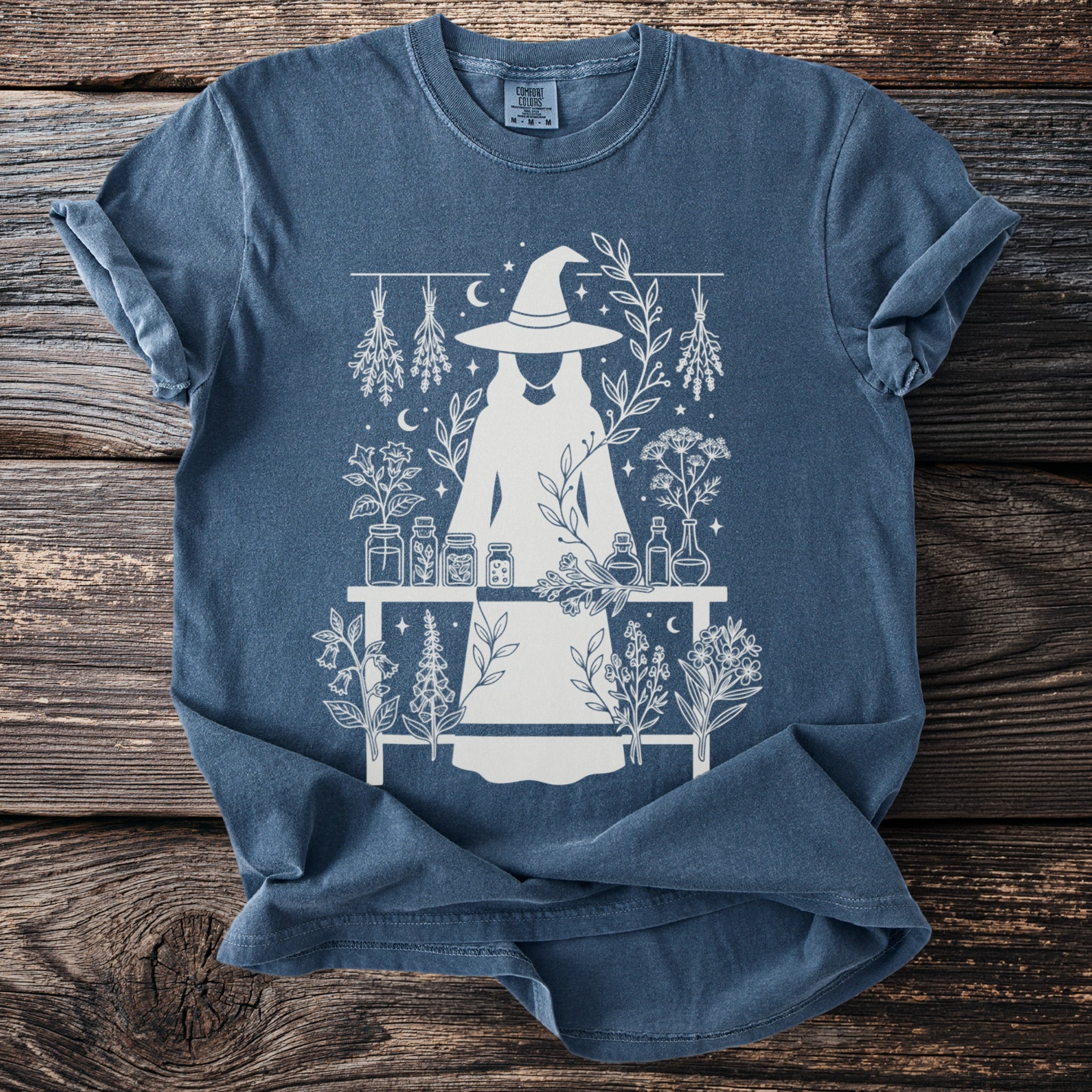 T-shirt with white graphic of a witch, potion bottles, herbs, and celestial elements.