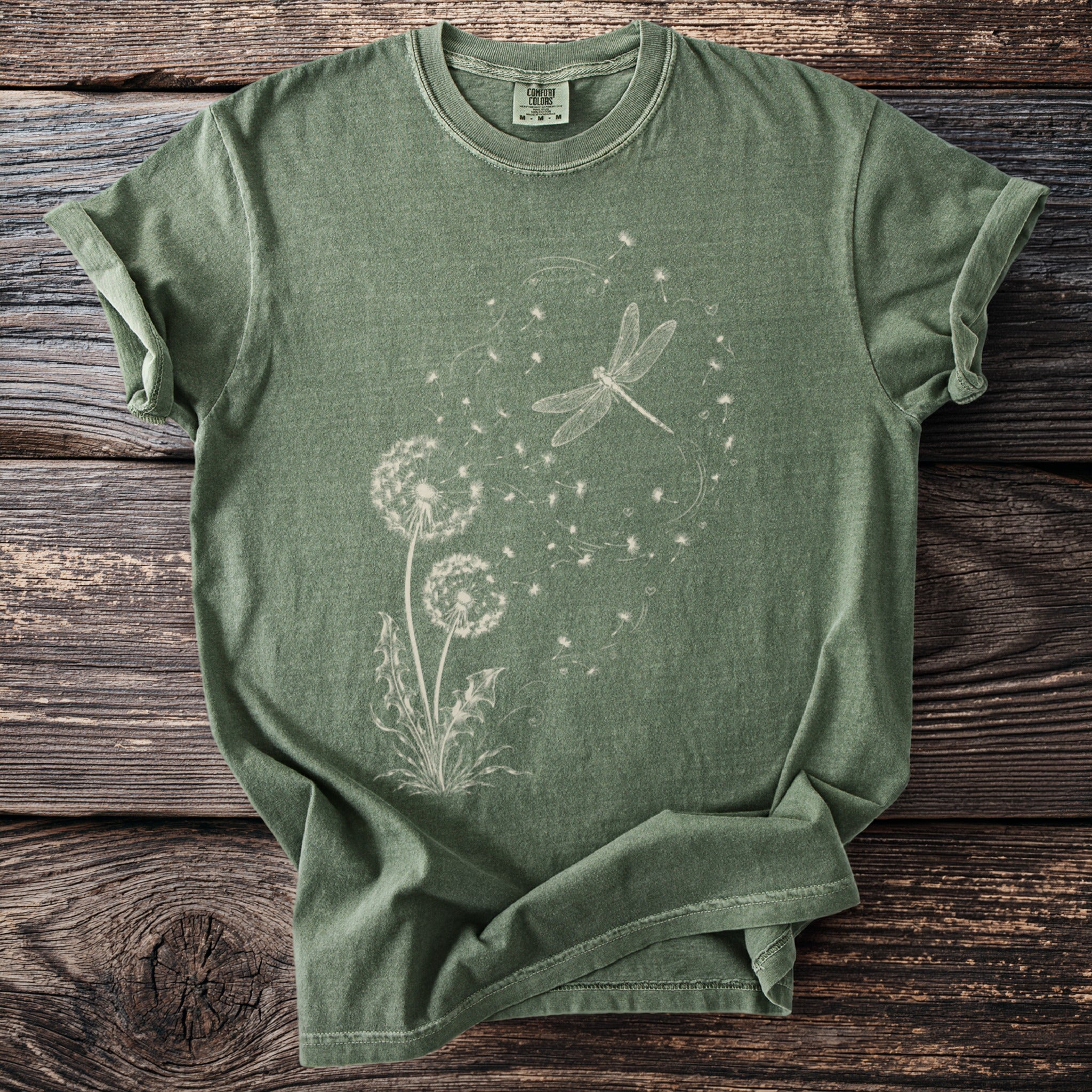 Green t-shirt with white dandelion and dragonfly graphic on a wooden background.