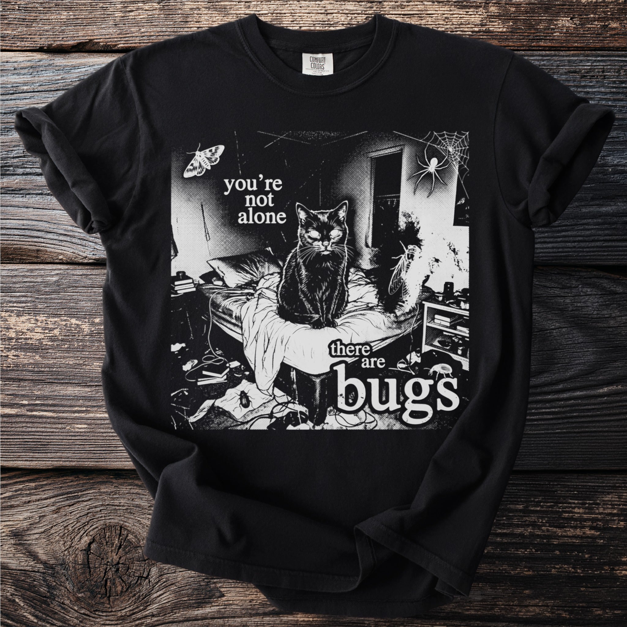 Black t-shirt with a cat on a bed surrounded by bugs and text saying "you're not alone there are bugs."
